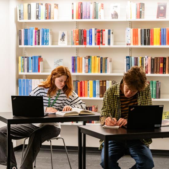 To unge sidder ved hvert deres bord og arbejder med bøger og computere. De sidder på et bibliotek. 